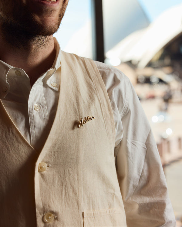 Man wearing a light-colored vest with a lapel pin, standing indoors with a blurred background of the Sydney Opera House.